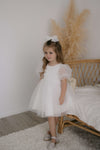 A young girl stands wearing our Gracie tulle puff sleeve flower girl dress in ivory.