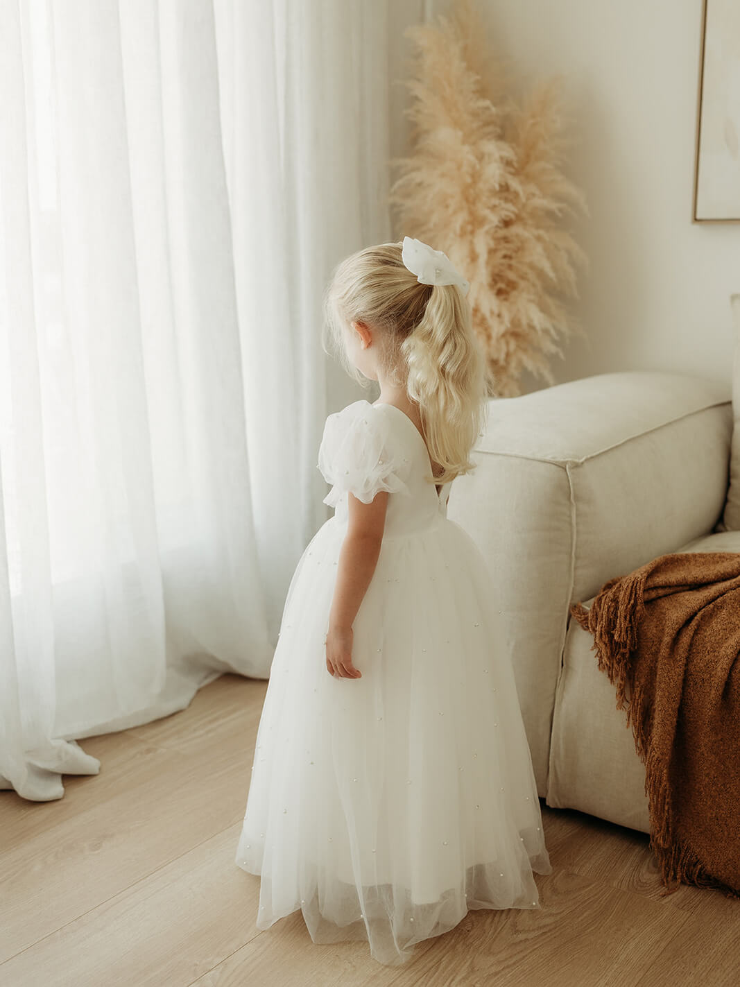 Gracie beaded tulle flower girl dress in ivory is worn by a young girl who stands near a window.
