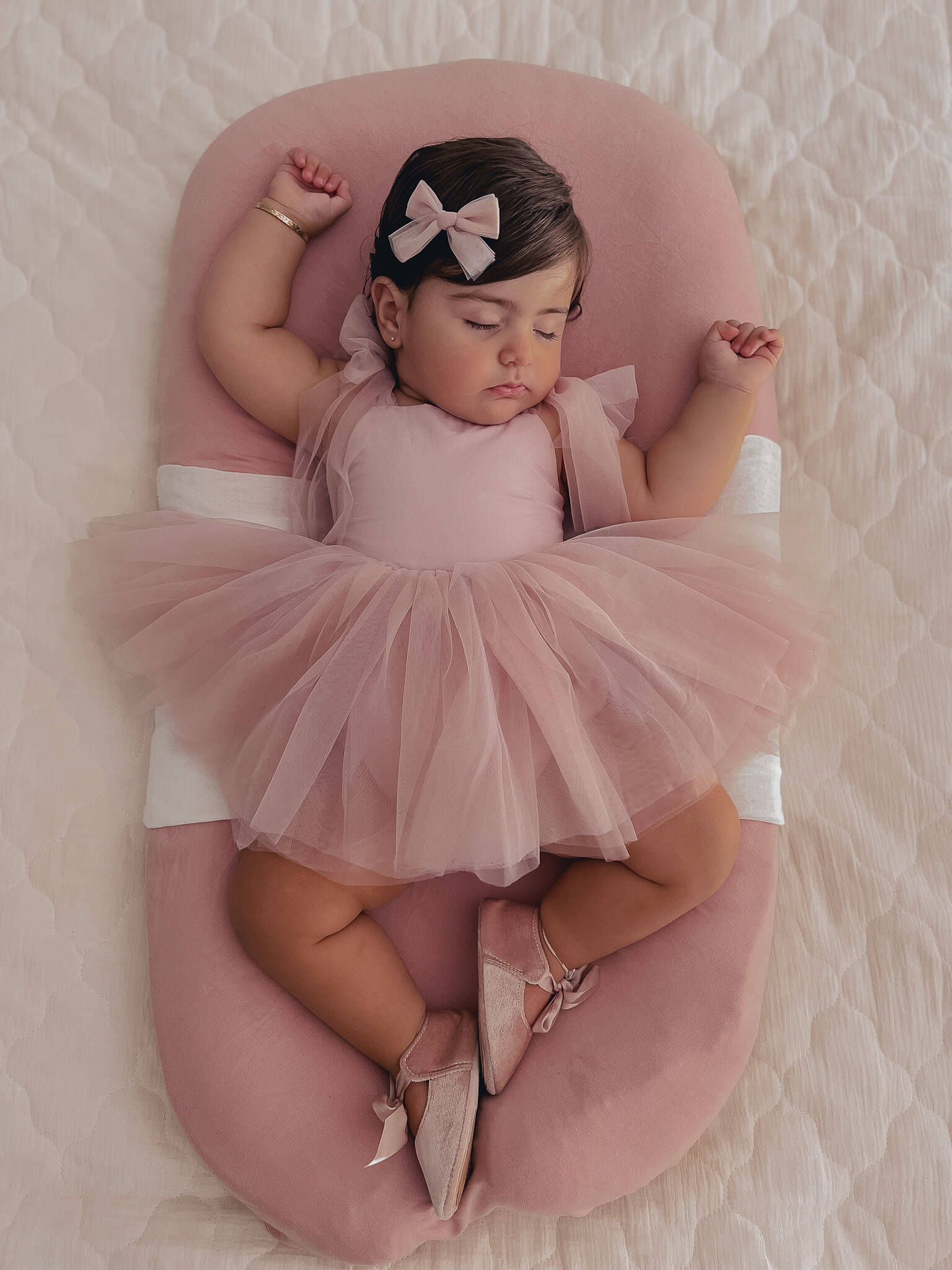 Harper baby flower girl romper in dusty pink is worn by a sleeping baby. She also wears a matching tulle bow clip in her hair.
