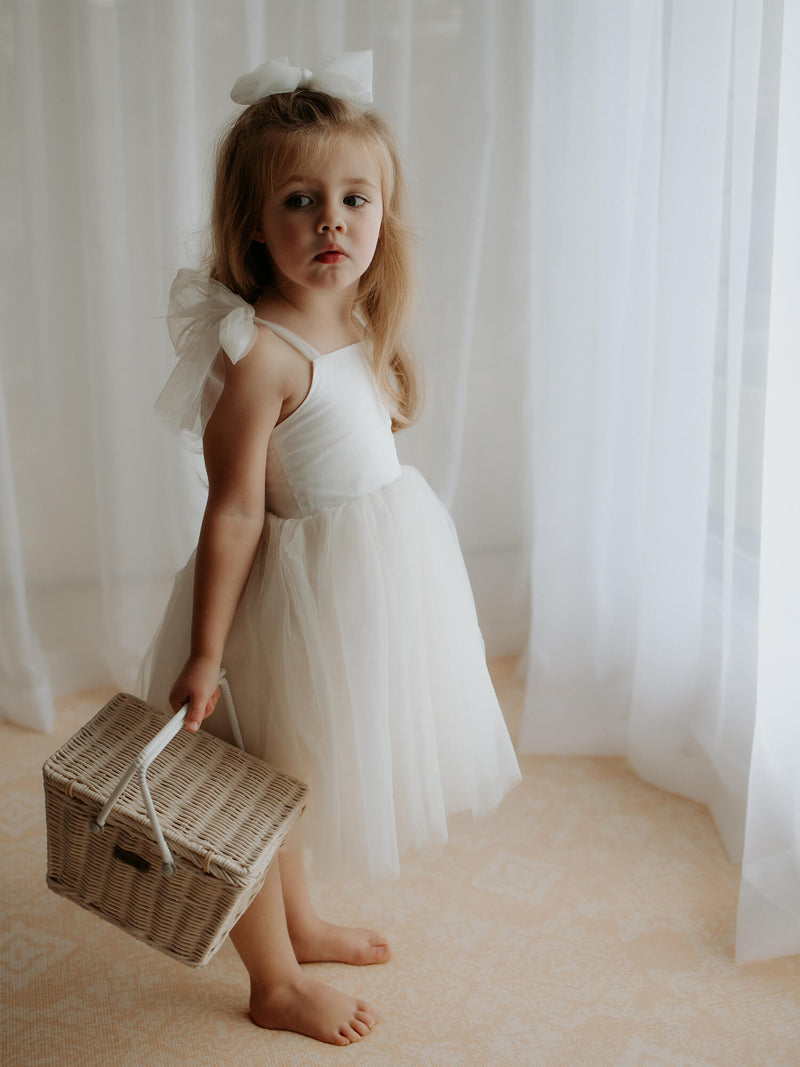 Harper tea length flower girl dress in cream, worn by a flower girl holding a basket. She also wears a cream tulle bow clip in her hair.
