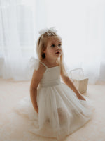A young girl sits wearing our Harper tea length flower girl dress in cream.