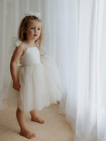 A girl stands by a white curtain at a window wearing our Harper tea length cream flower girl dress, with tulle tie straps.