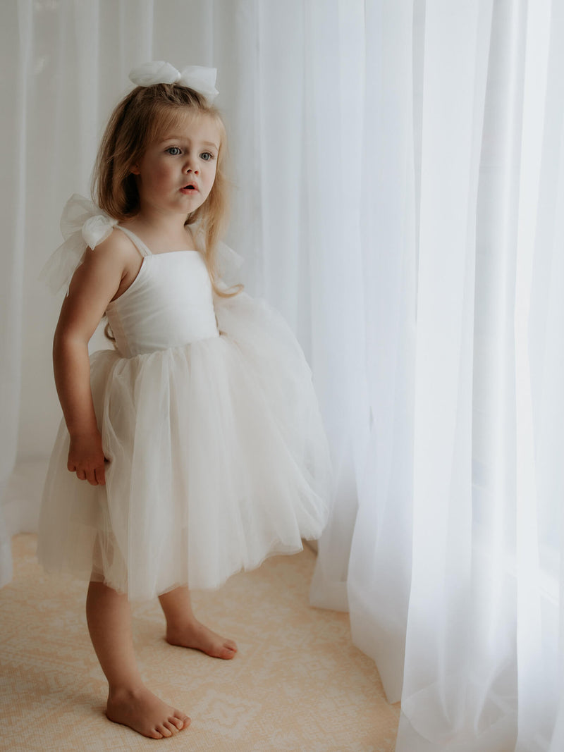 A girl stands by a white curtain at a window wearing our Harper tea length cream flower girl dress, with tulle tie straps.