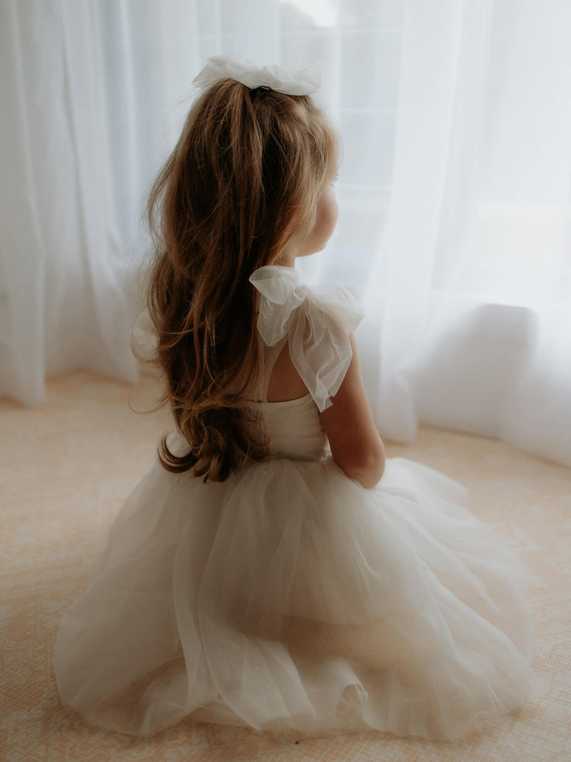 The back of our Harper tea length flower girl dress in cream, worn by a young girl. Showing the tulle tie straps, shirring at the back and soft tulle skirt.