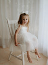 Harper cream flower girl dress is worn by a young girl who sits on a seat. She also wears a matching tulle bow in her hair.