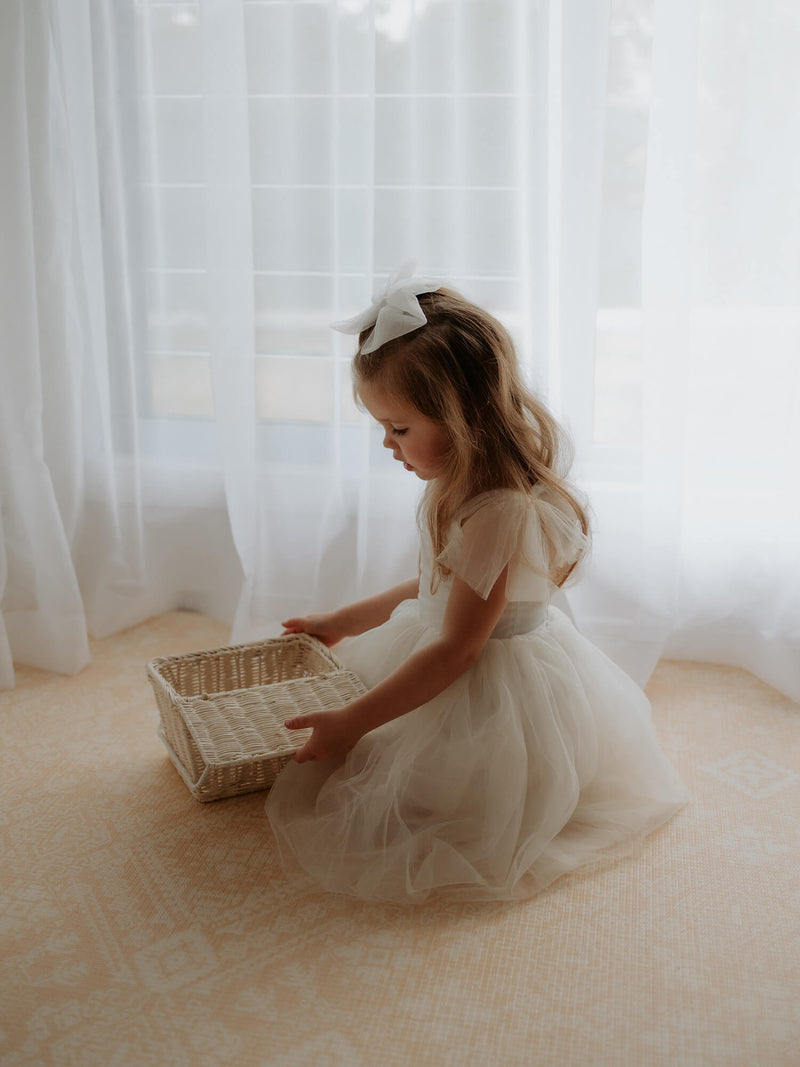 Cream flower girl dress, Harper tea length, is worn by a young girl who is looking into a basket. Showing the tulle tie straps and soft tulle skirt.