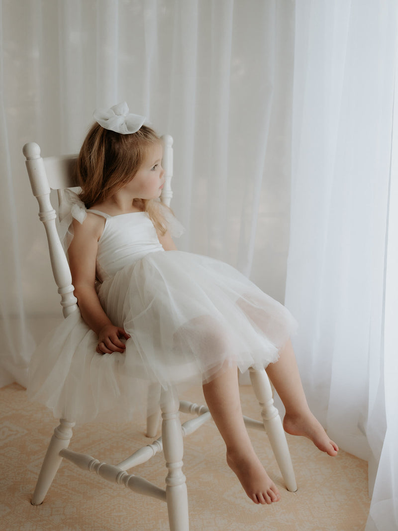 Harper cream flower girl dress with tulle tie straps is worn by a young girl, she also wears a matching tulle bow in her hair.