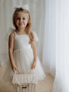 Harper cream flower girl dress is worn by a young girl who holds a basket. She also wears a matching cream tulle bow in her hair.