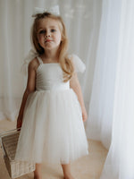 Harper tea length cream flower girl dress with tulle tie straps is worn by a flower girl with a tulle bow in her hair.