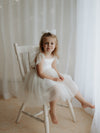 A young smiling flower girl sits on a seat wearing our Harper soft tulle flower girl dress in cream, along with a matching tulle bow hair clip.