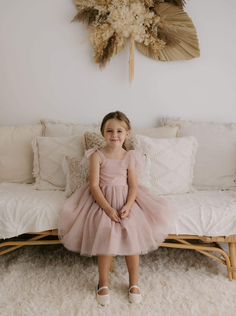 A little girl sits, smiling. She wears our Harper tea length dress in dusty pink, with tulle bow straps and a tea length soft tulle skirt.