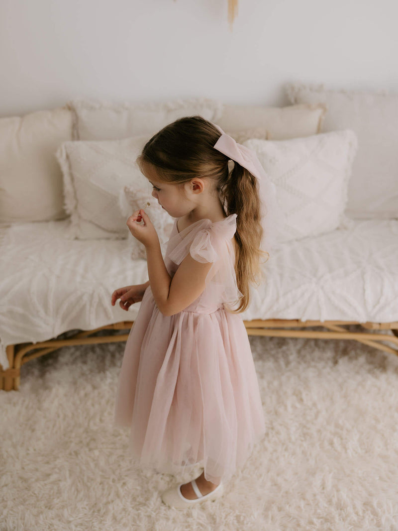 Harper tea length flower girl dress in dusty pink and matching large tulle bow in dusty pink, are worn by a young girl.