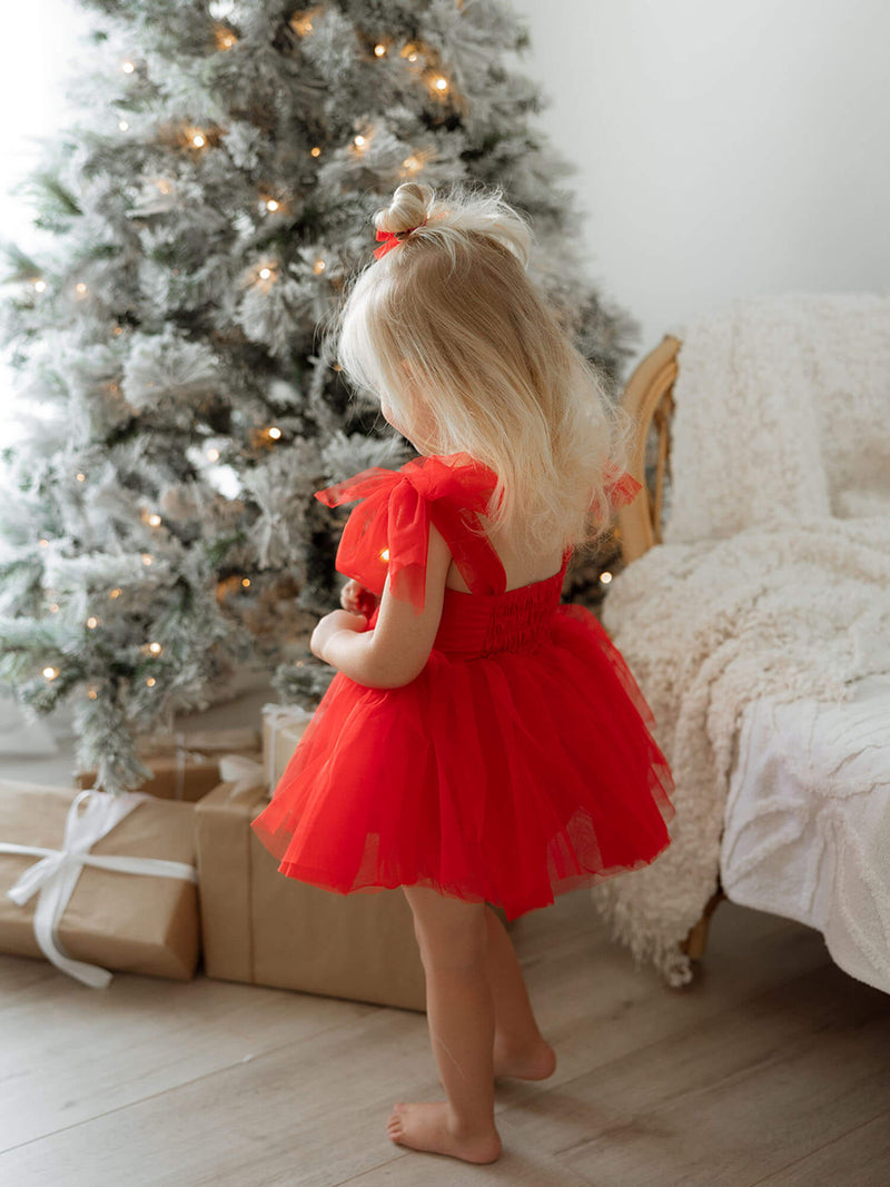 Harper ruby red Christmas baby romper is worn by a toddler who stands by a Christmas tree.