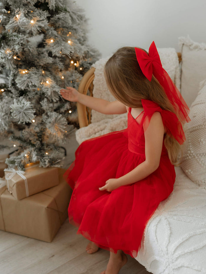 A young girl sits by a Christmas tree wearing our Harper ruby red bow sleeve dress and matching large ruby red tulle bow.