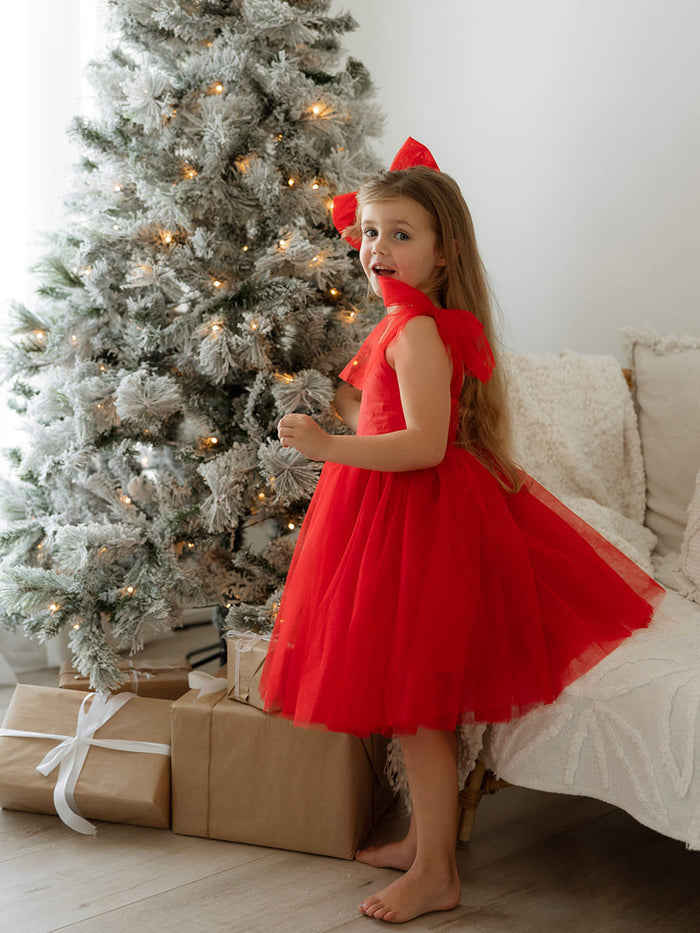 Harper ruby red tulle Christmas dress is worn by a young girl who stands by a Christmas tree.