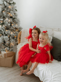 Sisters sit smiling by a Christmas tree, wearing matching ruby red dresses and tulle bows.