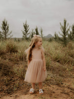 A young girl stands outside smiling, she wears our champagne Isla flower girl dress in tea length.