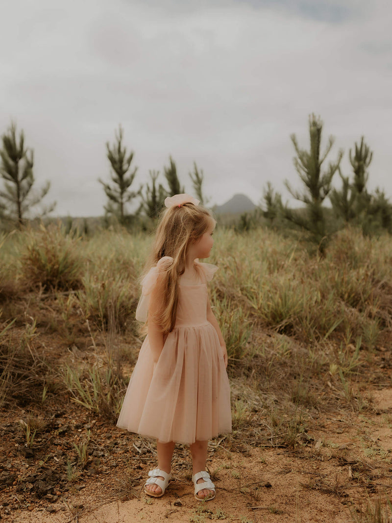A young girl wears our Isla champagne tulle bow sleeve flower girl dress and tulle bow hair clip in champagne.