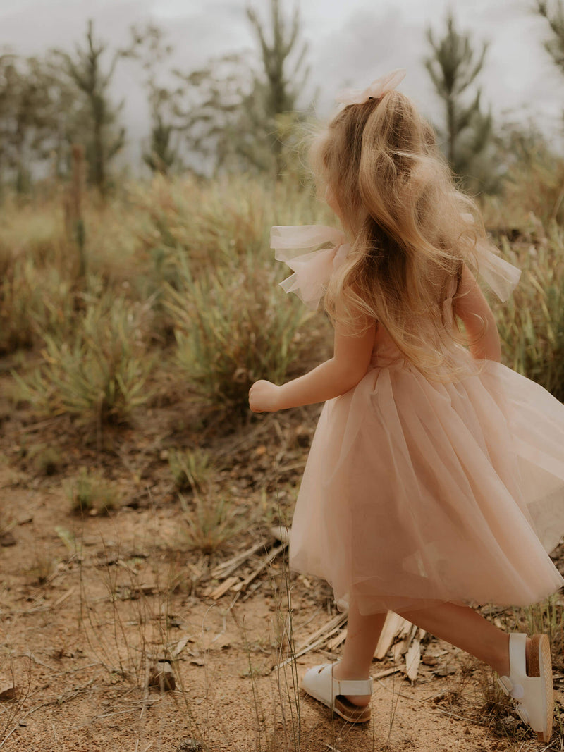 Young girl walks outside, wearing our Isla flower girl dress in champagne. Showing the soft tulle tie straps, and tea length champagne tulle skirt.