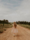 A young girl skips down a dirt road surrounded by pine trees, she wears our Isla champagne tulle flower girl dress.
