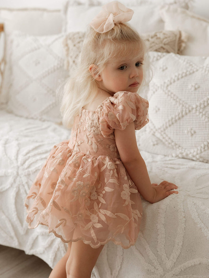 A toddler wears our Kenzi puff sleeve dusty rose baby flower girl romper and a matching tulle bow in her hair.