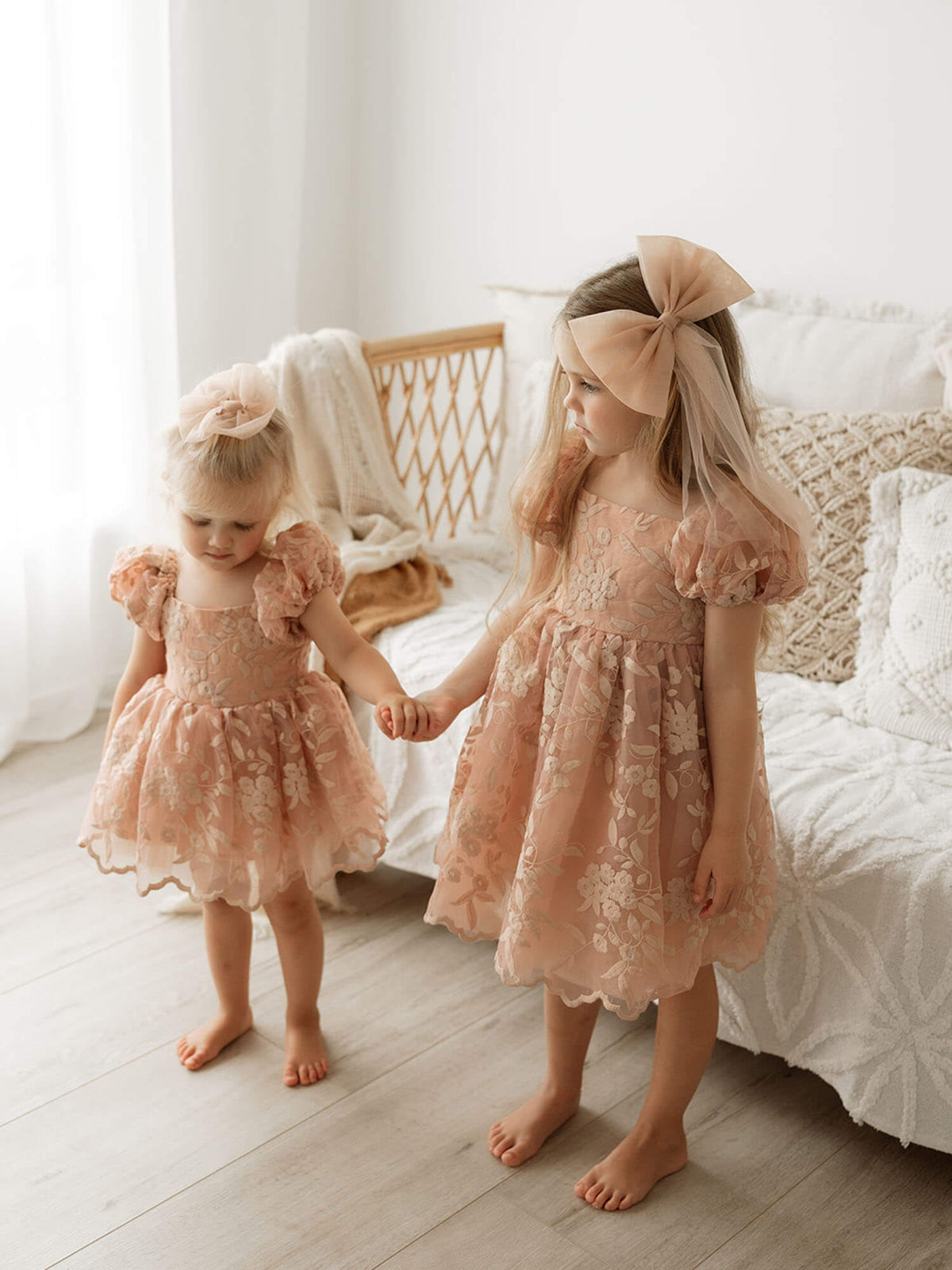 Sisters stand holding hands, they wear our Kenzi baby flower girl romper and Kenzi flower girl dress in dusty rose. They both also wear matching tulle bows in their hair.