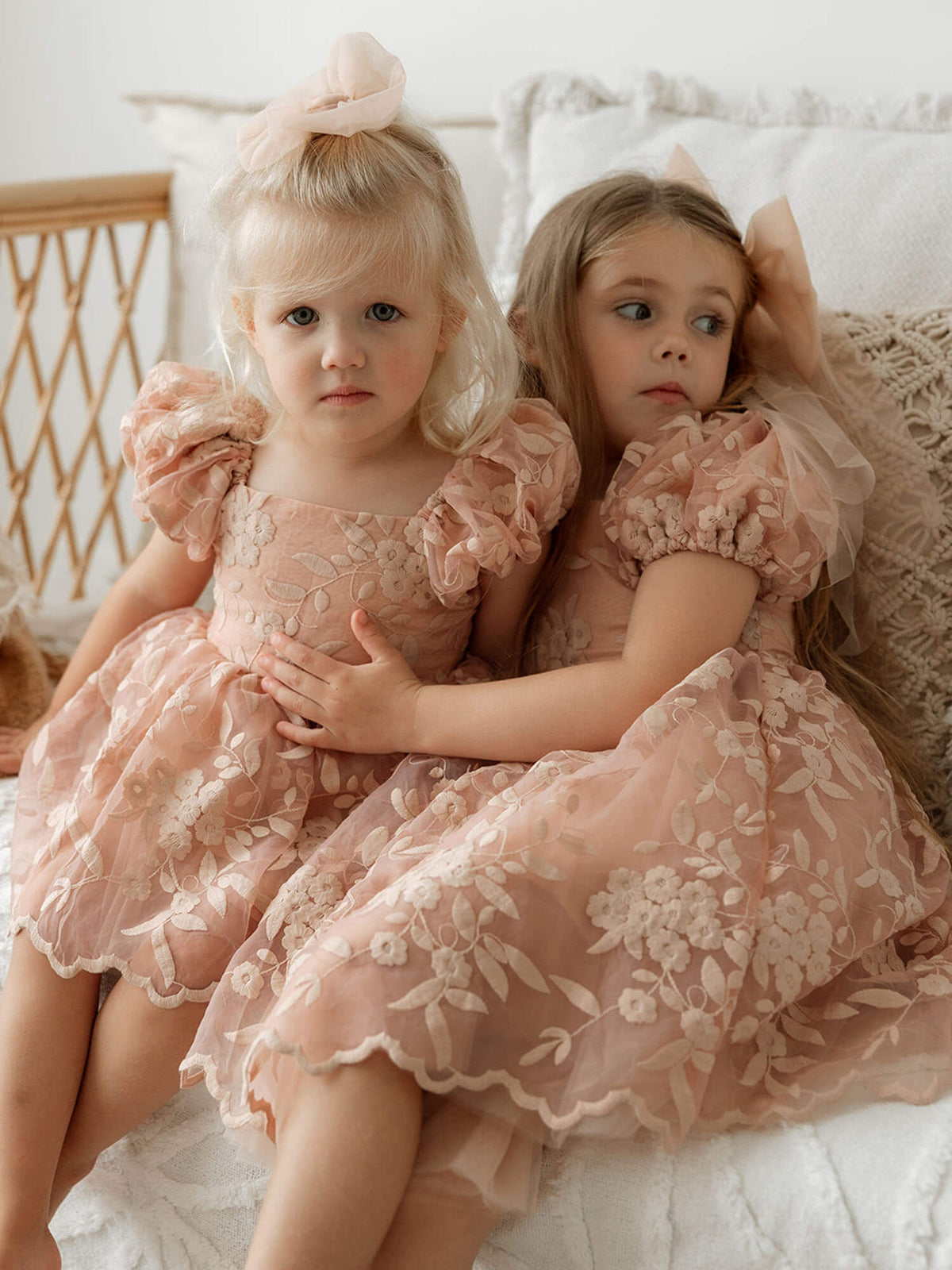 Two sisters sit beside each other wearing matching Kenzi dusty rose flower girl dress and Kenzi dusty rose baby flower girl dress, along with matching tulle bows in their hair.