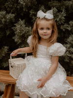 A young flower girl sits wearing our Kenzi lace puff sleeve flower girl dress in ivory, and medium tulle bow hair clip.