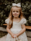 Kenzi puff sleeve flower girl dress and ivory tulle bow in medium are worn by a flower girl.