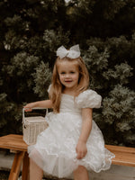 Lace puff sleeve flower girl dress is worn by a young girl who sits holding a flower girl basket.