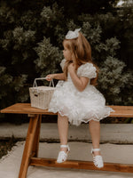A young girl sits on a stool wearing our Kenzi lace flower girl dress, holding a flower girl basket.