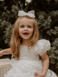A smiling flower girl wears our Kenzi lace puff sleeve flower girl dress and tulle bow in ivory.