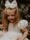 Kenzi puff sleeve lace flower girl dress is worn by a young girl, she also wears a medium tulle bow clip in ivory.