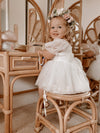 Toddler girl sits at a rattan table wearing our Layla baby flower girl romper in ivory and our Daisy flower crown.