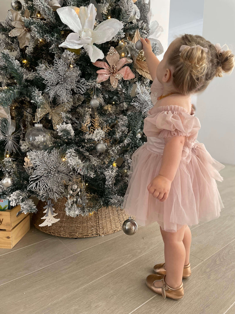 Layla baby dusty pink flower girl dress is worn by a young girl. She stands by a Christmas tree with blush and ivory decorations.