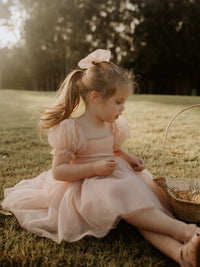 A young girl sits with a basket, she wears a tulle bow in her hair and our Champagne Layla flower girl dress.