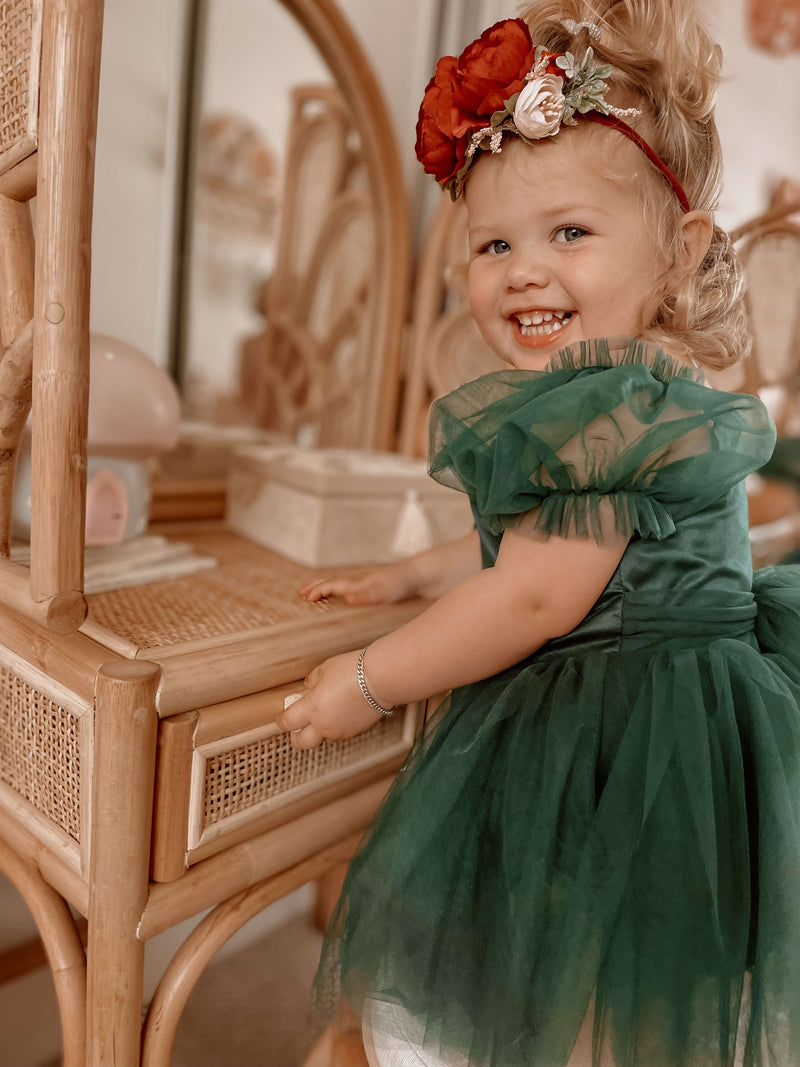 A toddler smiles at the camera wearing our Layla emerald green tulle Christmas romper, which can also be worn as a flower girl dress.