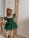 A toddler stands looking at a height chart, wearing our Layla emerald baby flower girl romper. Showing the back of the green baby romper with a tulle bow tied at the back.