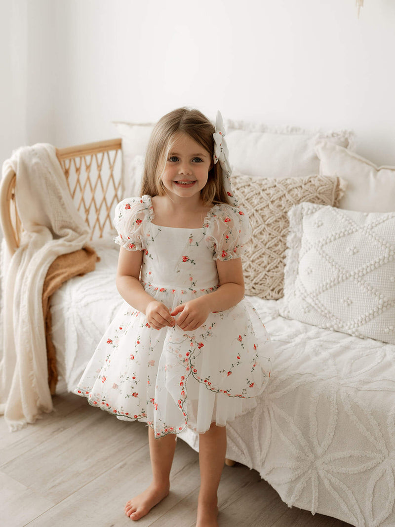 A little girl smiles wearing our red floral embroidered Layla dress and matching red floral hair bow.