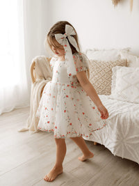 Layla organza floral embroidered dress is worn by a young girl. She wears a matching red floral embroidered bow in her hair.