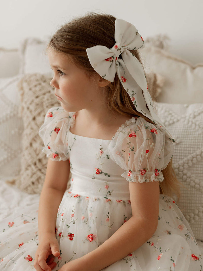 A little girl sits wearing a floral embroidered hair bow and matching floral embroidered Christmas dress.