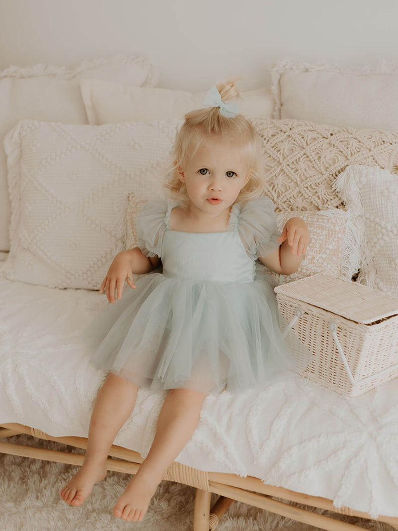 Layla dusty blue baby flower girl dress is worn by a young girl. She wears a tulle bow hairclip and sits on a chair with a flower girl basket.