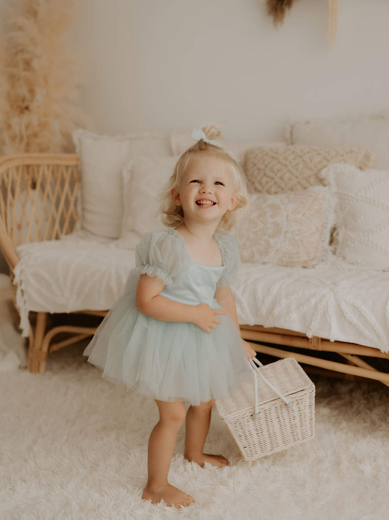 Layla dusty blue toddler flower girl romper is worn by a young girl. She stands holding a flower girl basket.