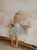 Layla dusty blue baby flower girl dress is worn by a young girl. She also wears a tulle pigtail bow in her hair.