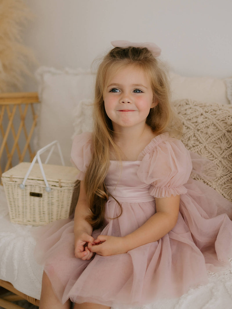 Tulle bow hair clip and tulle puff sleeve flower girl dress in dusty pink are worn by a young girl.