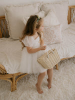 A young girl sits on a seat holding a basket, she wears our Layla puff sleeve flower girl dress and Eden flower crown.