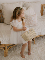 Layla flower girl dress in ivory is worn by a young flower girl who sits on a seat, holding a basket.