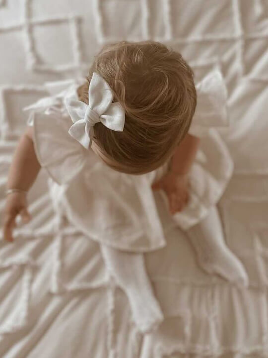 Baby sitting wearing an ivory dress and an ivory linen bow clip in her hair.