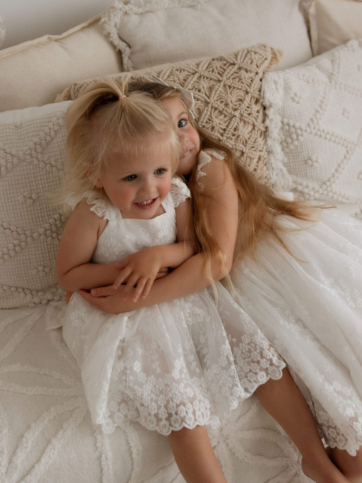 An older sister hugs her baby sister, who is wearing our Loulou ivory baby flower girl romper dress.