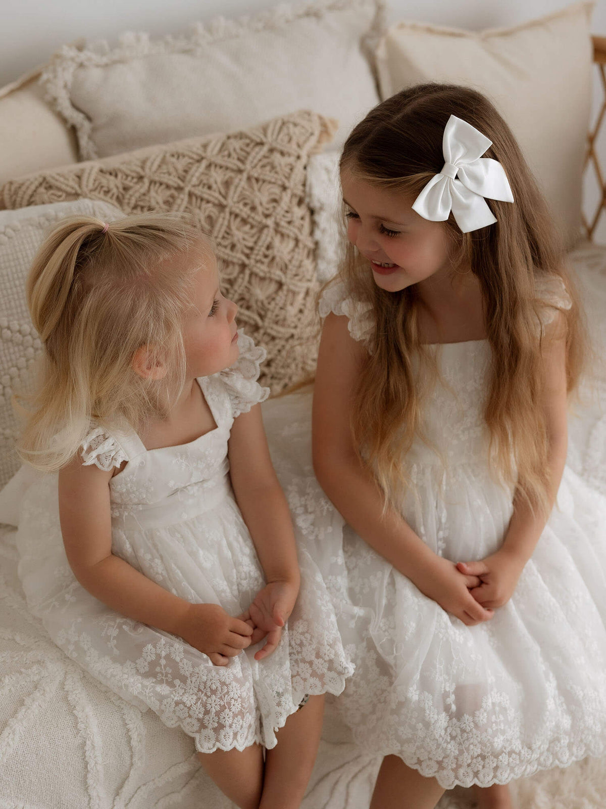 Sisters smile at each other while wearing matching Loulou toddler flower girl romper and Loulou flower girl dresses. A small ivory satin bow hair clip is also worn.